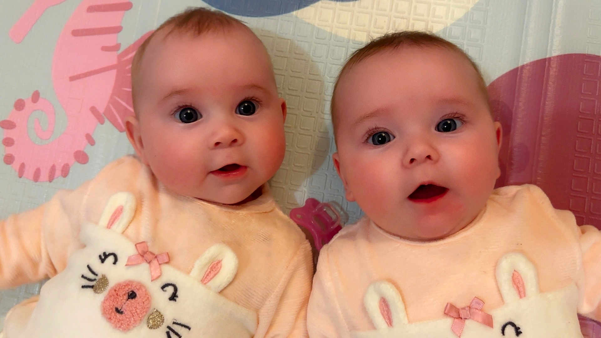 Romy Et Léna a rejoint le concours — aidez-le/la à gagner de superbes lots ! baby, infant, twin, cute, matching_clothes, bunny_design, peach_outfit, play_mat, pink_seahorse, wide_eyes, lying_down, indoor, child, adorable, face, expression, soft_lighting, closeup, two_babies, pastel_colors