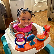 Blessing is registered to the contest to win money with this photo: baby, child, infant, walker, toy, pink_clothing, smiling, indoor, living_room, wooden_floor, hair, curly_hair, seat, play, colorful, furniture, cute, young_child, person, happy