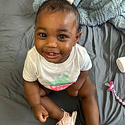 Gianna is registered to the contest to win money with this photo: baby, child, smiling, sitting, white_shirt, pink_sandals, hair_bow, bed, blanket, pacifier, happy, cute, infant, indoors, portrait, person, skin, footwear, toy, toddler
