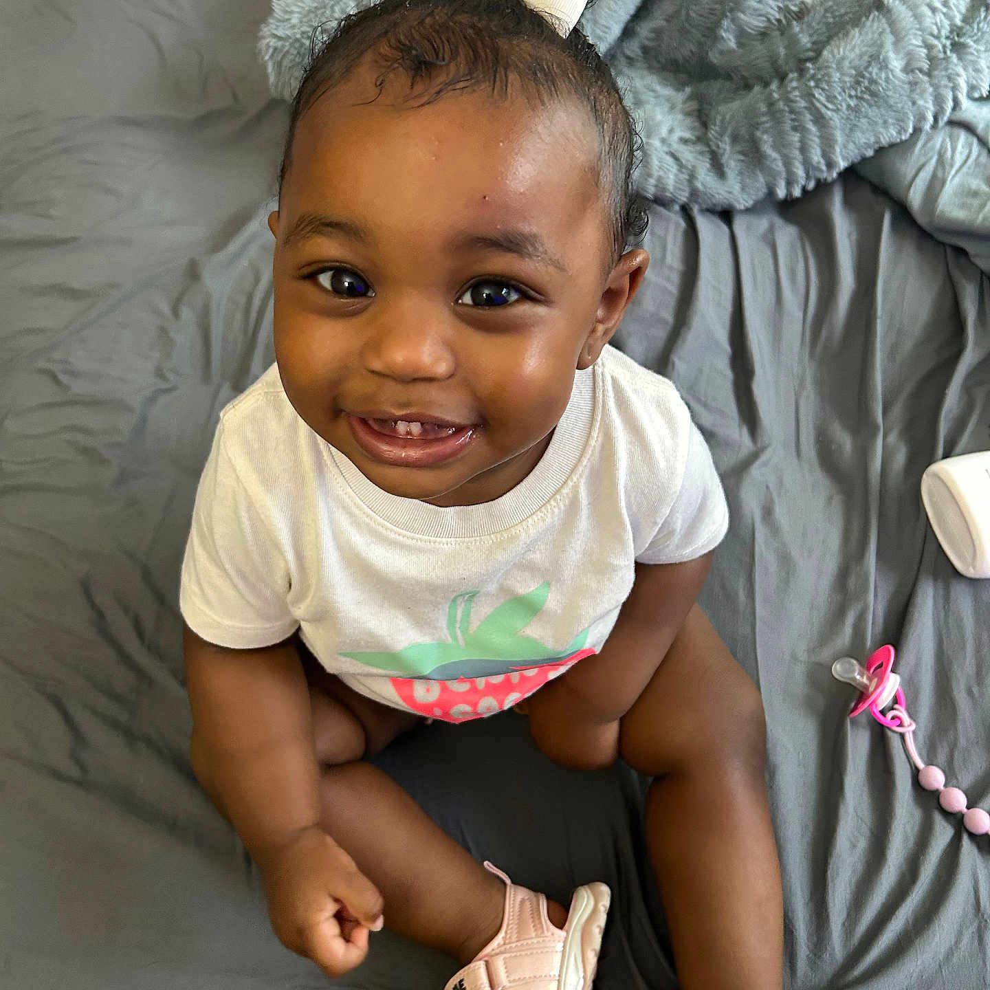 Gianna is registered to the contest to win money with this photo: baby, child, smiling, sitting, white_shirt, pink_sandals, hair_bow, bed, blanket, pacifier, happy, cute, infant, indoors, portrait, person, skin, footwear, toy, toddler