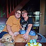 birthday, cake, candles, celebration, couple, dog, dogs, family, glasses, golden_retriever, home, indoors, living_room, man, portrait, puppy, seated, smiling, table, woman