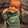 baby, infant, pacifier, onesie, clothing, striped_blanket, lying_down, hand, face, head, cute, newborn, child, indoors, text_on_clothing, raised_fist, blanket, soft_fabric, portrait, young_child