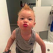 Xander is registered to the contest to win money with this photo: toddler, child, boy, hair_spikes, pucker, toy_car, bathroom, cabinet, open_door, gray_shirt, shorts, standing, playful, cute, expression, indoor, floor, white_wall, hand, face