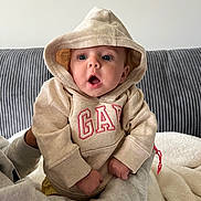 Danni is registered to the contest to win money with this photo: baby, hoodie, child, person, clothing, beige, indoor, couch, blanket, expression, surprised, cute, casual, sitting, warm, portrait, face, hands, soft, young