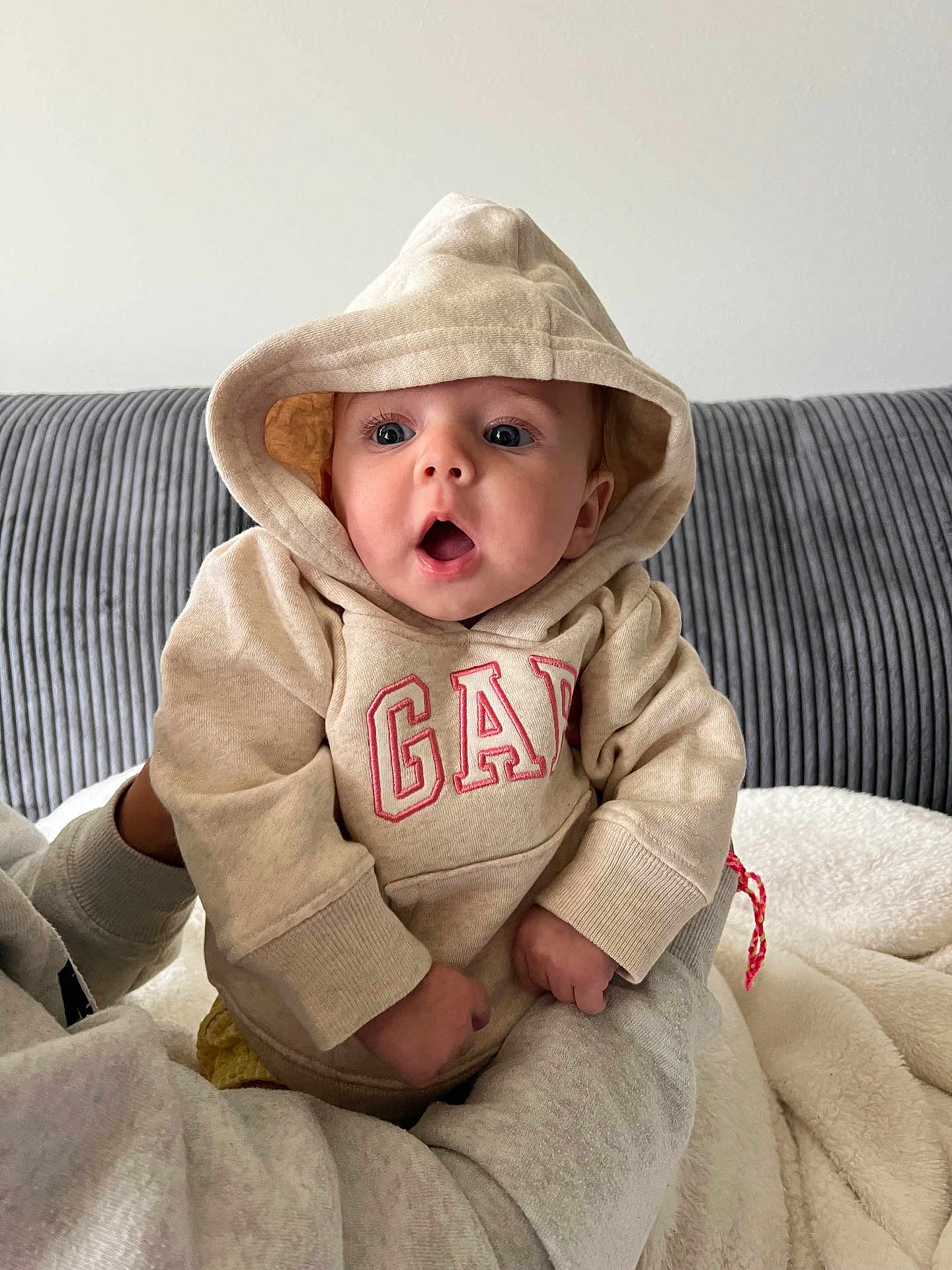 Danni is registered to the contest to win money with this photo: baby, hoodie, child, person, clothing, beige, indoor, couch, blanket, expression, surprised, cute, casual, sitting, warm, portrait, face, hands, soft, young