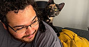 Hermione joined the competition — help win amazing prizes! man, cat, indoor, curly_hair, glasses, shoulder, tortoiseshell_cat, smile, decorations, pink_bows, heart_decoration, yellow_blanket, couch, pillow, facial_hair, casual_clothing, home, pet, cozy, curious