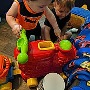 Ayedon is registered to the contest to win money with this photo: child, toddler, toy, play, indoor, floor, plastic_toy, colorful, vehicle_toy, phone_toy, blanket, pattern, wood_floor, furniture, curious, pointing, sitting, candid, young_children, interaction