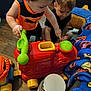 child, toddler, toy, play, indoor, floor, plastic_toy, colorful, vehicle_toy, phone_toy, blanket, pattern, wood_floor, furniture, curious, pointing, sitting, candid, young_children, interaction