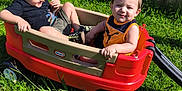 Ayedon is registered to the contest to win money with this photo: child, children, wagon, red_wagon, grass, outdoor, sunny, smile, happy, play, toy, fence, greenery, summer, casual_clothing, shoes, two_kids, sitting, daylight, fun
