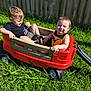 child, children, wagon, red_wagon, grass, outdoor, sunny, smile, happy, play, toy, fence, greenery, summer, casual_clothing, shoes, two_kids, sitting, daylight, fun