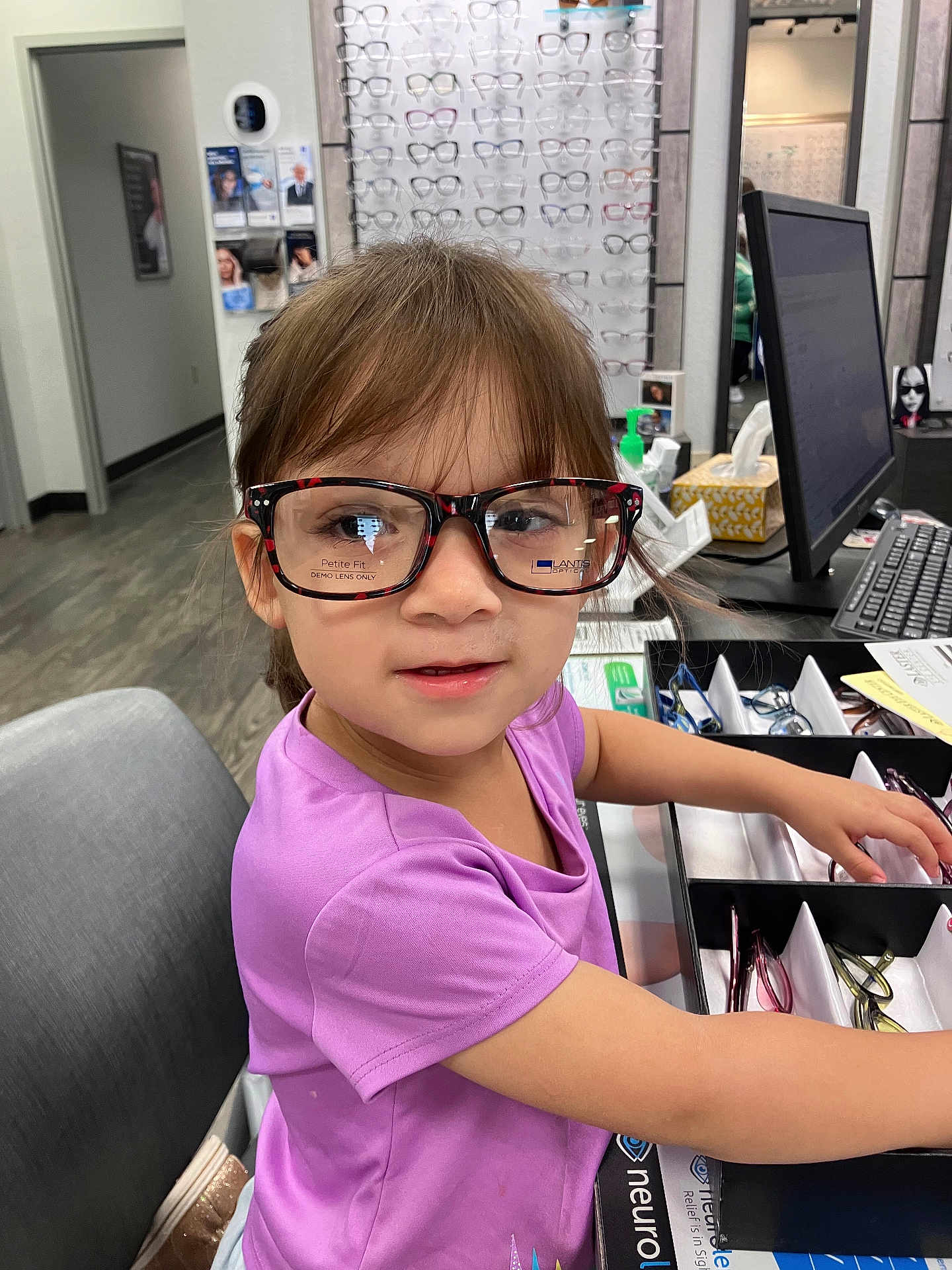 Athena is registered to the contest to win money with this photo: child, glasses, optical_store, purple_shirt, tray, face, person, indoor, eyewear, curly_hair, desk, chair, computer_monitor, keyboard, display, shelf, floor, hand, reflection, young