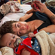 Aries is registered to the contest to win money with this photo: baby, adult, blanket, bed, pacifier, curly_hair, tattoo, phone, hand, red_cloth, blue_clothing, indoor, relaxing, person, face, skin, lying_down, comfort, cozy, sleepwear