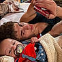 Aries is registered to the contest to win money with this photo: baby, young_man, blanket, pacifier, bed, phone, hand, face, curly_hair, tattoo, clothing, red, blue, indoor, relaxing, portrait, comfort, family, lying_down, candid