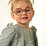 child, toddler, glasses, pigtails, green_dress, portrait, smile, cute, fashion, indoor, young_child, person, studio, buttoned_dress, face, happy, eyewear, hair, light_background, sitting