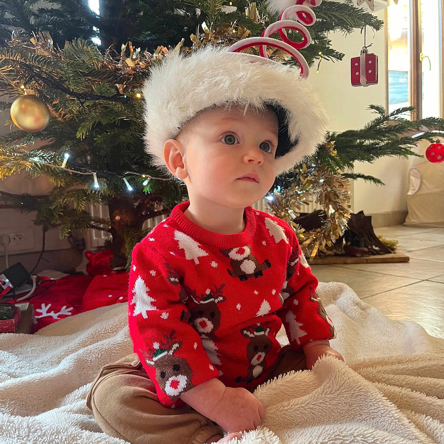 Raphaël a rejoint le concours — aidez-le/la à gagner de superbes lots ! blanket, child, christmas_tree, cozy, cute, decorations, festive, floor, holiday, holiday_hat, indoors, lights, ornaments, portrait, red_sweater, reindeer_pattern, seasonal, sitting, toddler, young_child