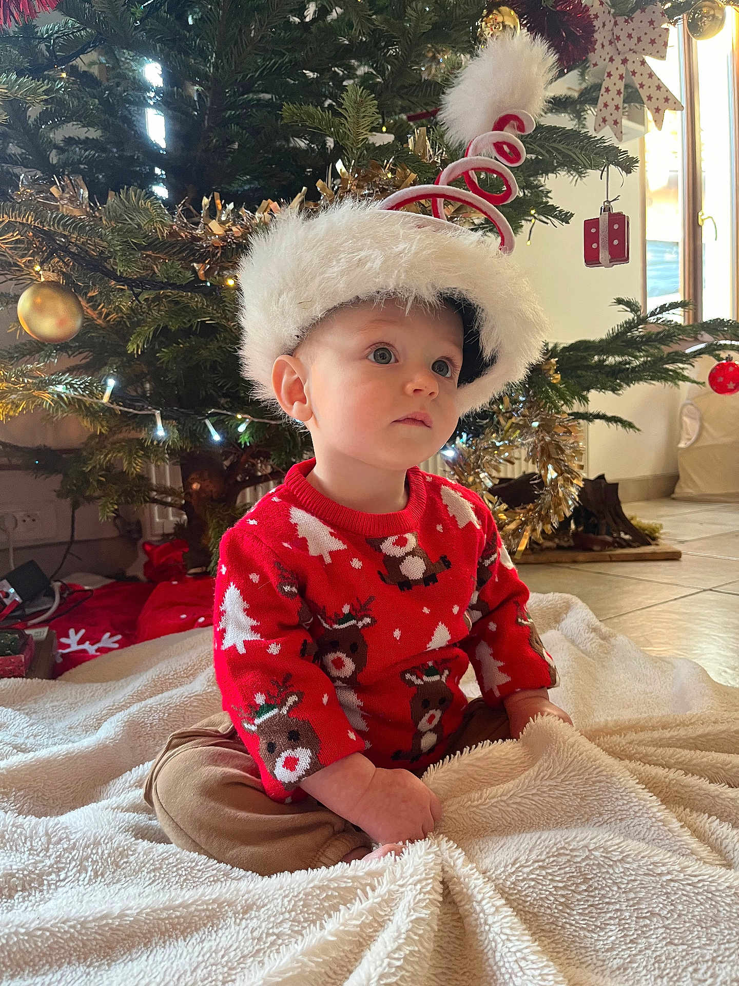 Raphaël a rejoint le concours — aidez-le/la à gagner de superbes lots ! toddler, child, sitting, christmas_tree, holiday_hat, red_sweater, reindeer_pattern, blanket, indoors, decorations, festive, cute, cozy, floor, lights, ornaments, holiday, seasonal, portrait, young_child
