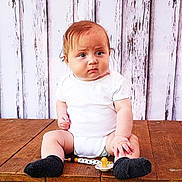 Reggie is registered to the contest to win money with this photo: baby, infant, child, white_onesie, black_socks, pacifier, wooden_surface, wooden_background, beaded_clip, serious_expression, sitting, cute, portrait, indoors, plush, human, young_child, casual_clothing, floor, hands