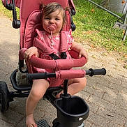 Iris participe au concours pour gagner de l'argent avec cette photo : toddler, child, stroller, tricycle, pink, pacifier, pigtails, outdoor, daylight, park, greenery, signpost, footwear, pavement, smiling, cute, baby, seatbelt, toy, handlebars