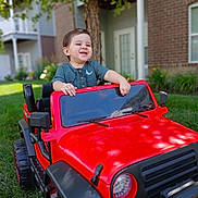 Marcelo is registered to the contest to win money with this photo: backyard, casual_clothing, child, daylight, door, fun, grass, greenery, happy, house, outdoor, play, portrait, red, smiling, toddler, toy_car, tree, vehicle, window