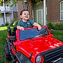 child, toddler, toy_car, red, outdoor, grass, smiling, happy, greenery, vehicle, play, casual_clothing, daylight, tree, backyard, house, window, door, fun, portrait