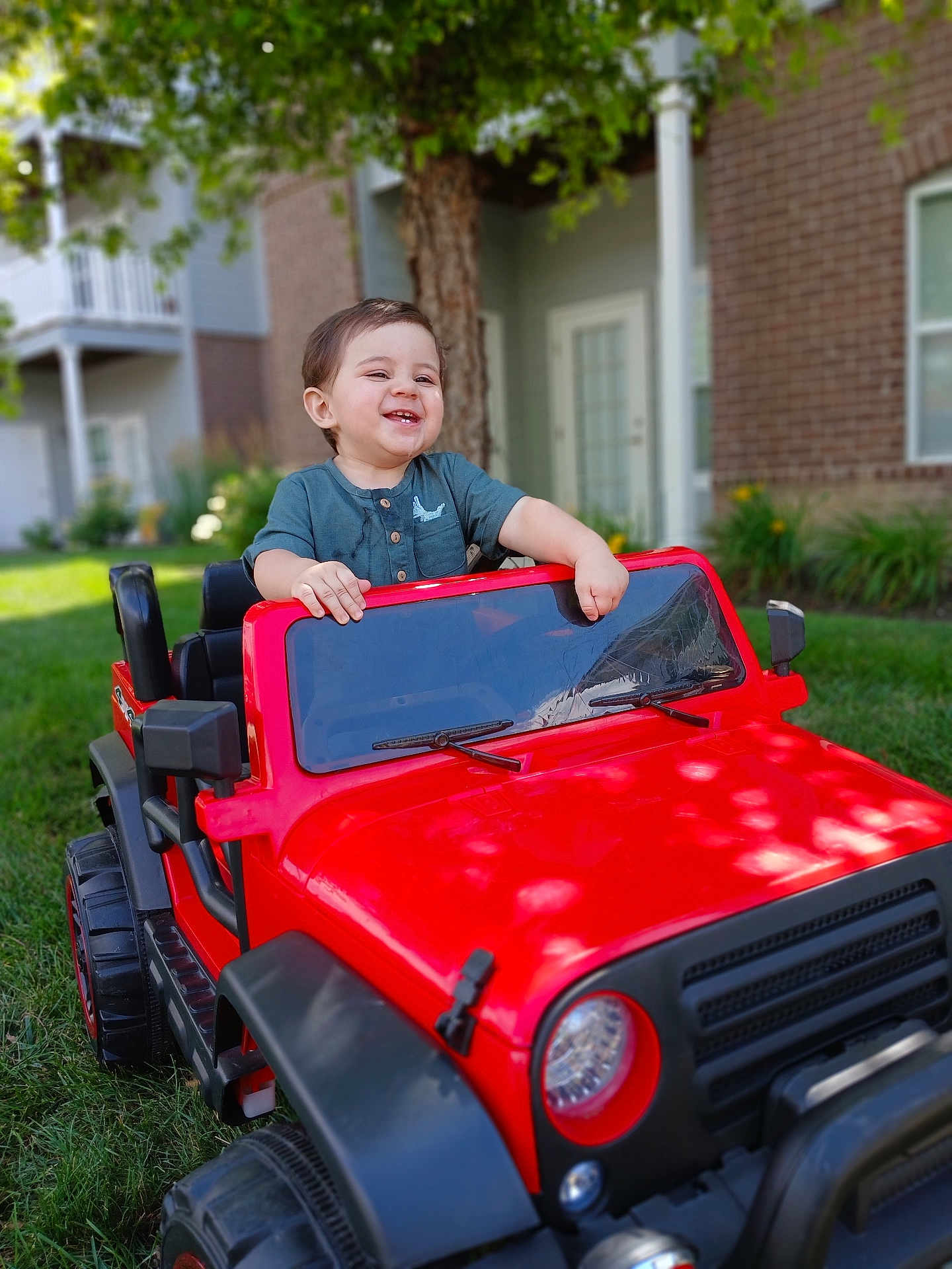 Marcelo is registered to the contest to win money with this photo: child, toddler, toy_car, red, outdoor, grass, smiling, happy, greenery, vehicle, play, casual_clothing, daylight, tree, backyard, house, window, door, fun, portrait