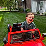 Marcelo is registered to the contest to win money with this photo: child, toddler, toy_car, red_car, outdoor, grass, greenery, smiling, happy, summer, play, fun, vehicle, young_child, casual_clothing, residential_area, daylight, person, parked_toy, nature