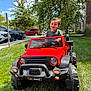child, toddler, toy_car, red, grass, outdoor, sunny, smiling, greenery, tree, suburban, car, vehicle, play, fun, daytime, happy, person, jeep, sidewalk