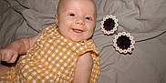 Nova is registered to the contest to win money with this photo: baby, smiling, infant, yellow_dress, gingham, blanket, sunglasses, daisy, flower, cute, happy, lying_down, child, portrait, face, skin, arm, clothing, indoor, soft