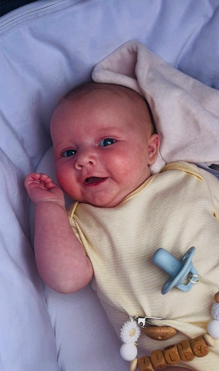 Tilly joined the competition — help win amazing prizes! baby, infant, smile, face, hand, blanket, pacifier, clothing, beads, name_tag, cute, lying_down, soft_texture, happy, skin, blue_eyes, yellow_outfit, closeup, indoors, child
