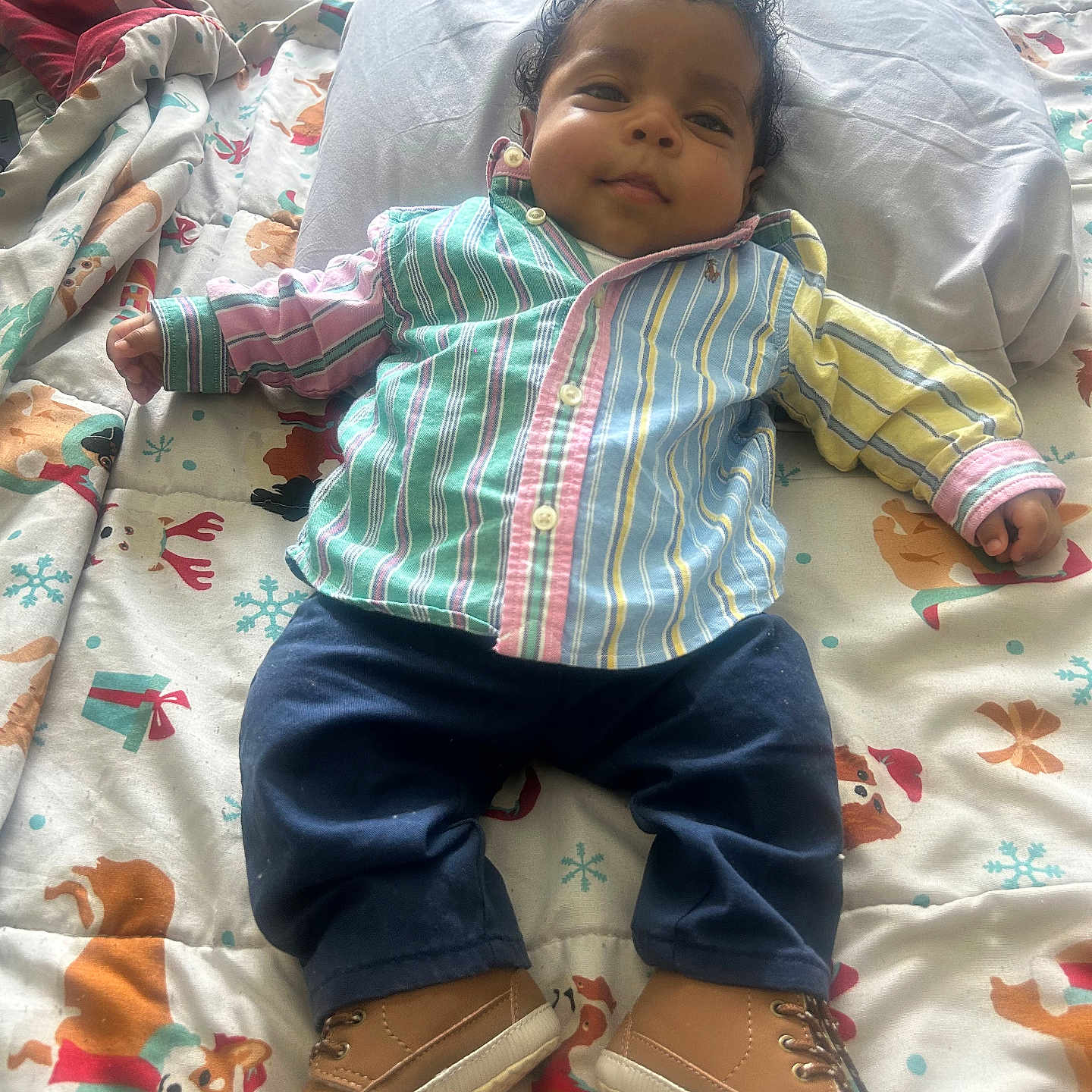 Jai Mari joined the competition — help win amazing prizes! baby, child, infant, striped_shirt, navy_pants, tan_shoes, curly_hair, bed, blanket, holiday_theme, reindeer_pattern, pillow, lying_down, cute, smiling, indoors, cozy, colorful, person, portrait