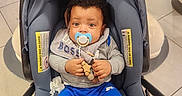 Prince joined the competition — help win amazing prizes! baby, infant, car_seat, pacifier, toy, blue_pants, grey_bib, curly_hair, indoor, floor_tiles, child, person, clothing, seat_belt, footwear, cute, sitting, looking_at_camera, holding, comfort