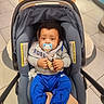 baby, infant, car_seat, pacifier, toy, blue_pants, grey_bib, curly_hair, indoor, floor_tiles, child, person, clothing, seat_belt, footwear, cute, sitting, looking_at_camera, holding, comfort