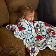 Scarlett Drew joined the competition — help win amazing prizes! baby, blanket, brown_couch, child, pacifier, indoor, seated, portrait, cozy, wrapped, floral_pattern, soft_texture, young_child, curly_hair, comfortable, expression, looking_away, home, furniture, casual