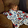 baby, blanket, brown_couch, child, pacifier, indoor, seated, portrait, cozy, wrapped, floral_pattern, soft_texture, young_child, curly_hair, comfortable, expression, looking_away, home, furniture, casual
