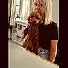 blonde_hair, bookshelf, casual_clothing, cozy, curtains, cute, dog, friendly, furniture, home, indoor, light, pet, portrait, purple_harness, relaxed, smiling, table, window, woman