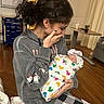 Valentina Jade. joined the competition — help win amazing prizes! baby, blanket, cup, curly_hair, cushion, duck_pattern, emotional, hat, holding, hoodie, hospital_room, indoor, newborn, person, plaid, portrait, seated, table, woman, wood_floor