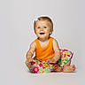baby, child, toddler, portrait, smiling, happy, sitting, orange_shirt, floral_skirt, hair_bow, blonde_hair, barefoot, studio, white_background, colorful, fashion, infant, face, indoor, photography