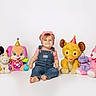 Lilah is registered to the contest to win money with this photo: child, toddler, baby, portrait, sitting, smiling, birthday, party_hat, stuffed_animals, plush_toys, mickey_mouse, teddy_bear, lion, unicorn, pink_bear, denim_overalls, bow_headband, indoor, white_background, colorful