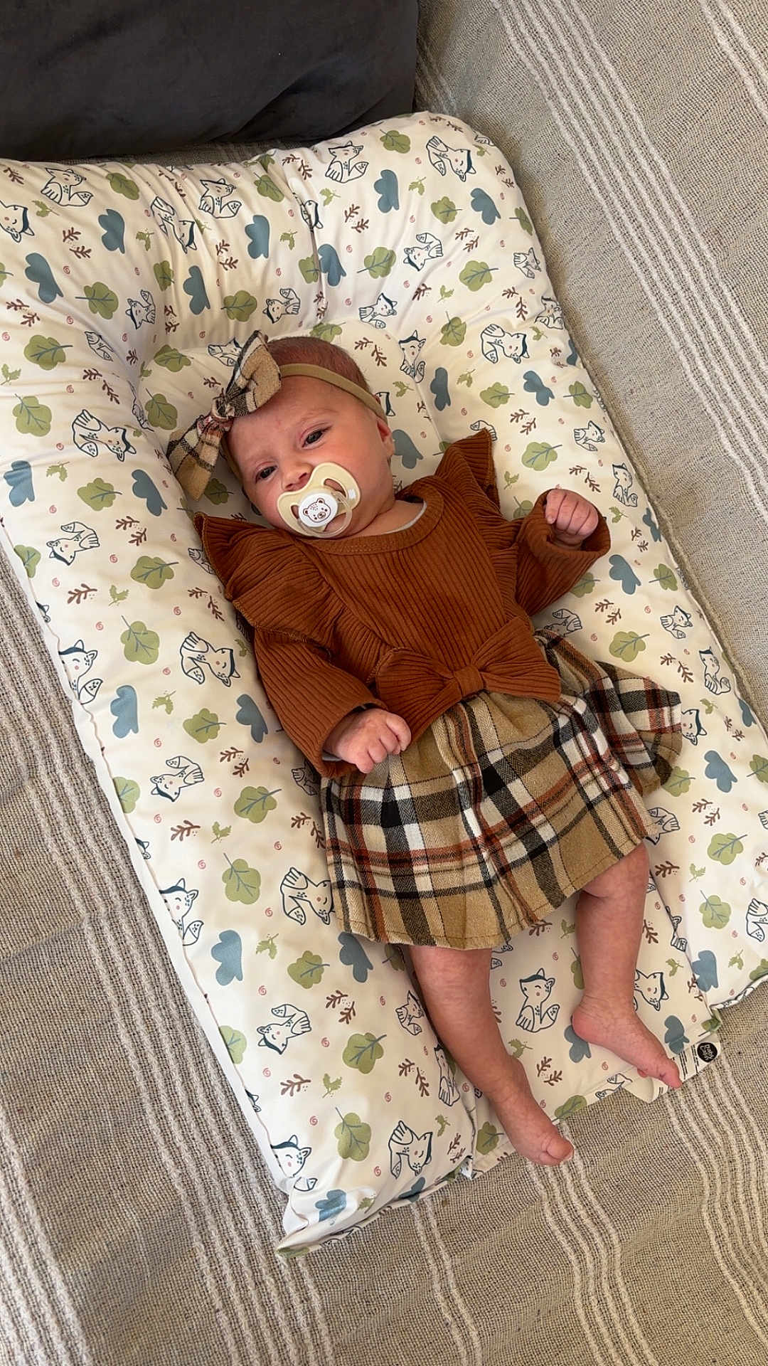 Maëlie a rejoint le concours — aidez-le/la à gagner de superbes lots ! baby, infant, pacifier, headband, bow, plaid_skirt, brown_top, cushion, fox_pattern, leaves_pattern, striped_rug, indoor, child, cute, soft_lighting, relaxed, lying_down, clothing, fabric, decor