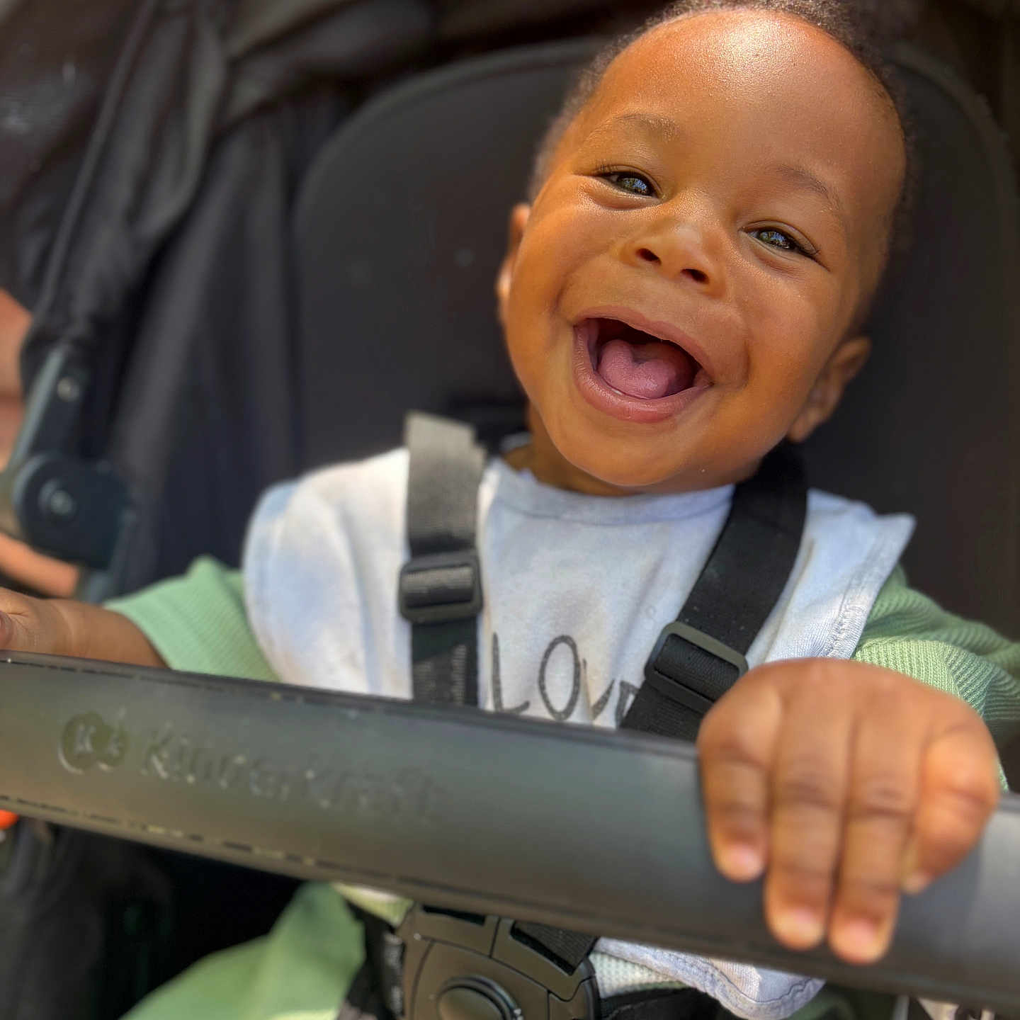 Mayvin participe au concours pour gagner de l'argent avec cette photo : baby, baby_bib, black_hair, child, closeup, clothing, cute, daylight, face, green_sleeves, hand, happy, infant, outdoor, person, portrait, seatbelt, smiling, stroller, toddler