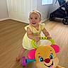 baby, child, indoor, toy, walker, yellow_dress, headband, flooring, smile, stroller, playful, happy, person, floor, window, door, bright, colorful, cute, standing