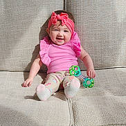 Evelyn is registered to the contest to win money with this photo: baby, baby_clothes, beige_couch, child, colorful_toy, couch, cute, feet, happy, headwear, indoor, infant, legs, pink_headband, pink_top, playful, sitting, smiling, soft_lighting, toy