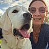 dog, golden_retriever, person, woman, smiling, sunglasses, outdoor, sky, clouds, tongue_out, pet, happy, friendship, portrait, face, necklace, blue_shirt, daylight, nature, closeup