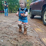 Lakelynn is registered to the contest to win money with this photo: child, toddler, mud, boots, denim, overalls, outdoor, dirt_road, car, woman, trees, grass, nature, daylight, casual_clothing, smiling, playing, person, vehicle, background