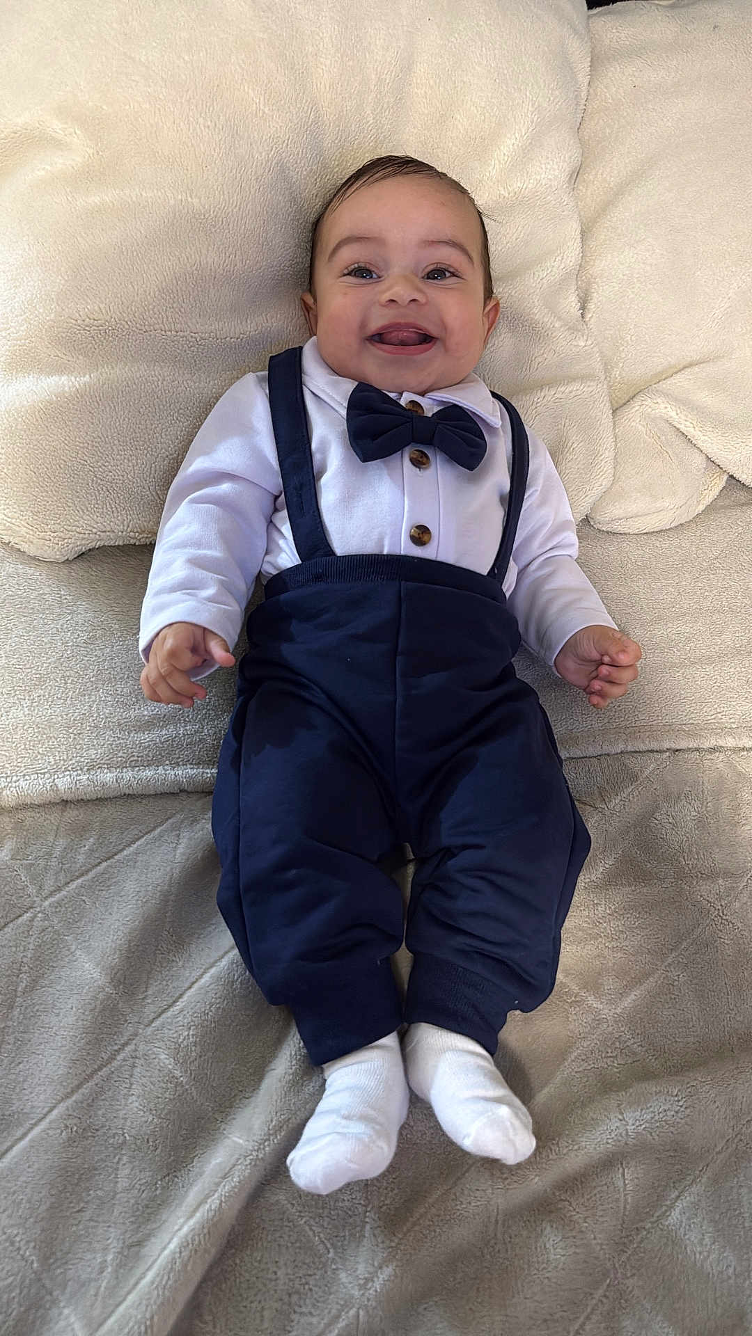 Yumel is registered to the contest to win money with this photo: baby, smiling, bow_tie, suspenders, white_shirt, pants, socks, blanket, couch, happy, child, portrait, indoor, cute, infant, clothing, face, hands, feet, lying_down