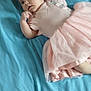 baby, infant, pink_dress, blue_bedspread, bed, lying_down, curious, finger_near_mouth, soft_fabric, indoor, child, person, young, cute, small, toddler, blanket, colorful, head, face