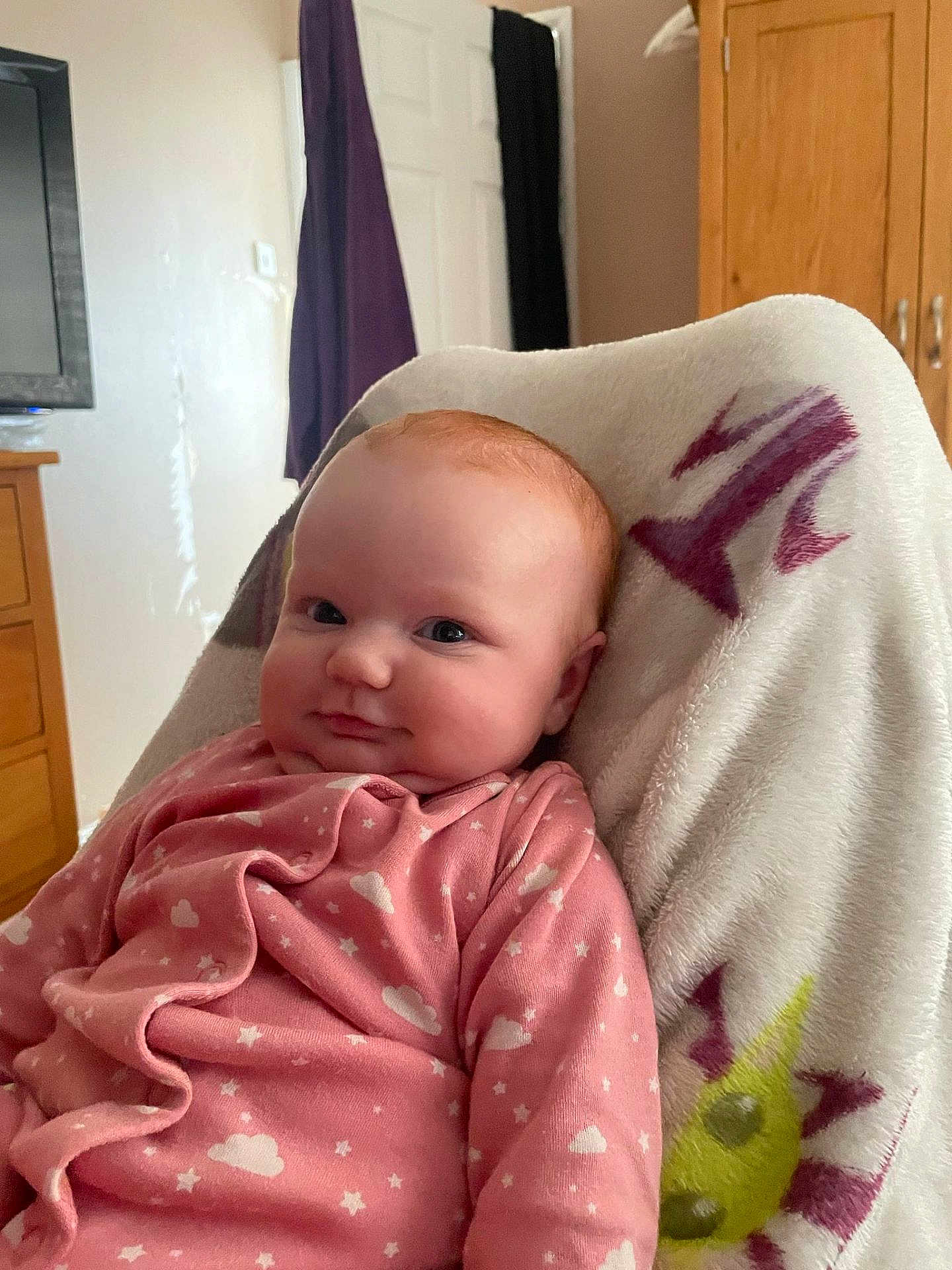 Elora is registered to the contest to win money with this photo: baby, infant, pink_clothing, blanket, indoor, face, child, cute, soft_texture, furniture, wooden_drawer, door, curtain, smile, head, portrait, cozy, warm, sleepwear, eyes