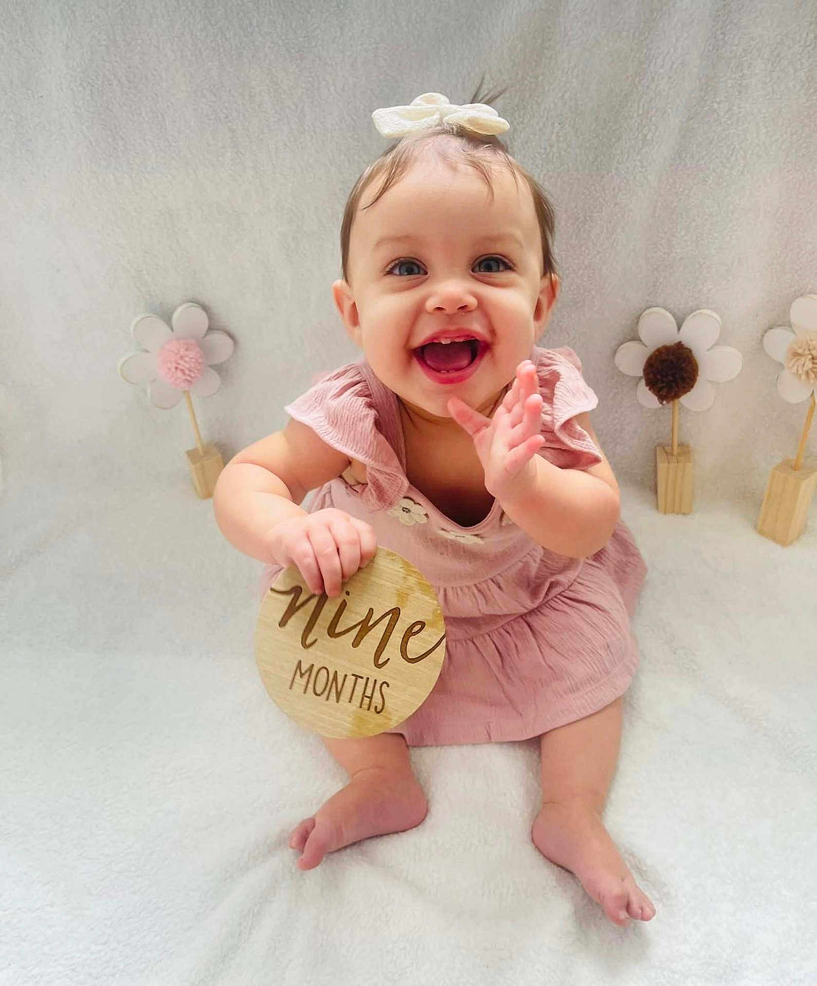 Meadow is registered to the contest to win money with this photo: baby, child, infant, girl, smiling, clapping, pink_dress, bow, wooden_sign, nine_months, sitting, white_blanket, decorations, flowers, happy, cute, portrait, indoors, soft_lighting, celebration