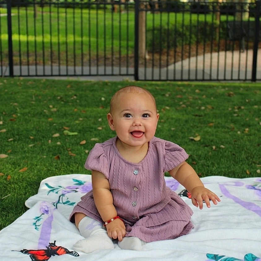Lucia is registered to the contest to win money with this photo: baby, infant, child, smiling, purple_dress, blanket, grass, outdoor, park, fence, happy, cute, sitting, nature, daylight, people, playful, young, portrait, lawn