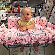 Lucia is registered to the contest to win money with this photo: baby, shopping_cart, pink_cushion, polka_dot, warehouse, store, aisle, paper_products, smiling, child, infant, barefoot, happy, face, cute, retail, cart, floor, clothing, person